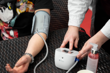 Mężczyzna ma założony mankiet ciśnieniomierza na przedramię, a obok stojąca osoba uruchamia elektroniczny aparat do pomiaru ciśnienia krwi.