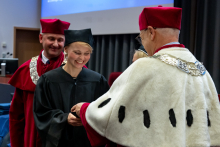 Kobieta w czarnej todze odbiera dokument z rąk rektora. Uśmiecha się.