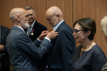 Rektor przypina odznaczenie innemu mężczyźnie podczas uroczystości. Obok stoją trzy osoby i obserwują sytuację. Wszyscy mają eleganckie stroje. 