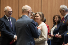 Rektor przypina odznaczenie kobiecie i rozmawia z nią. Kilka osób stoi obok i czeka na swoją kolej. Wszyscy mają eleganckie stroje i zachowują poważny, skupiony wyraz twarzy. Atmosfera jest uroczysta.