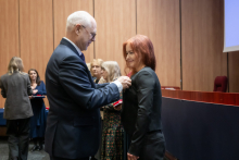 Rektor przypina odznaczenie kobiecie podczas uroczystości. Kobieta stoi naprzeciwko niego i patrzy na niego poważnie. W tle kilka osób przygotowuje kolejne wyróżnienia. Atmosfera jest spokojna i uroczysta.