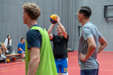 Mężczyzna w czarnej koszulce unosi piłkę do koszykówki nad głową, przygotowując się do rzutu na hali sportowej. Obok niego stoją dwaj inni mężczyźni obserwujący akcję, a w tle widać osoby siedzące na ławce oraz fragment boiska.