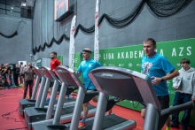 Mężczyźni biegną na bieżniach ustawionych w rzędzie podczas wydarzenia sportowego na hali. Obok stoi kobieta obsługująca stanowisk