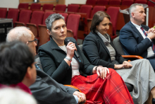 Osoba zabierająca głos podczas panelu dyskusyjnego, trzyma mikrofon i siedzi w rzędzie foteli wraz z innymi uczestnikami wydarzenia.