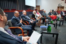 Panel dyskusyjny w auli; uczestnicy siedzą w półokręgu na fotelach, jeden z nich trzyma notatki, a na stolikach przed nimi stoją butelki z wodą.