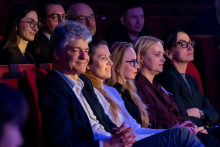 Pięć osób siedzi w pierwszym rzędzie sali i klaszcze po zakończonym występie. Wszyscy kierują wzrok w stronę sceny. Ich postawa jest swobodna, a wyraz twarzy sugeruje życzliwe skupienie. W tle widać kolejnych widzów, którzy również obserwują wydarzenie.