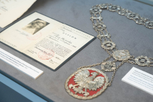 Gablotowa ekspozycja przedstawiająca historyczny dyplom oraz ceremonialny łańcuch z dużym emblematem orła, związane z działalnością Wydziału Farmaceutycznego.