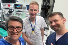 Trzech mężczyzn. Ubrani są w fartuchy lekarskie w różnych kolorach. Za nimi widać sprzęt medyczny. Uśmiechają się.