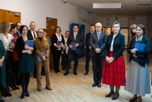 Grupa osób stojąca w korytarzu podczas uroczystego spotkania; uczestnicy trzymają teczki i słuchają przemówienia