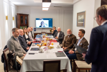 Grupa elegancko ubranych osób siedzi przy stole w czasie spotkania. Na ścianie widać ekran telewizyjny. Jeden mężczyzna, odwrócony tyłem do zdjęcia, przemawia. 