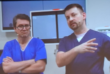 Dwóch mężczyzn w fartuchach medycznych stoi na sali zabiegowej, w tle widać sprzęty medyczne.