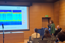 Kilka osób w sali konferencyjnej. Jedna z nich stoi z mikrofonem w ręce i zadaje pytanie. W tle widać ekran multimedialny z wyświetlaną prezentacją.