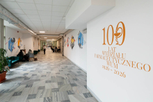 Grupa kilku osób siedzi przy małym stoliku w korytarzu uczelni, pracując wspólnie nad dokumentami. Na ścianie za nimi widoczne są kolorowe grafiki przedstawiające historyczne wydarzenia związane z wydziałem.