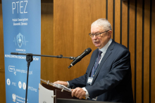 Mężczyzna w garniturze stoi przy mównicy i prowadzi wystąpienie. Trzyma notatki i mówi do mikrofonu. Obok niego stoi roll‑up z informacjami o konferencji.