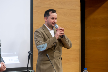 Mężczyzna w brązowej marynarce stoi przy mównicy i przemawia do publiczności, trzymając mikrofon w dłoniach. Obok niego widoczny jest fragment ekranu prezentacji