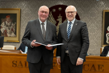 Dwóch mężczyzn stoi obok siebie, są elegancko ubrani. Jeden z nich trzyma teczkę z dokumentem. W tle siedzą dwie osoby oraz widać duże godło z orłem. 