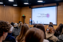 Prelegent stojący przed publicznością prowadzi wykład na tle dużego ekranu z prezentacją o roli mediów społecznościowych w nauce i praktyce, w nowoczesnej sali wykładowej