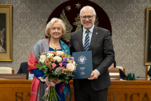 Kobieta i mężczyzna stoją obok siebie, są elegancko ubrani. Kobieta trzyma duży bukiet kwiatów. W tle siedzą dwie osoby oraz widać duże godło z orłem. 