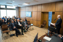 Sala w czasie konferencji. Kilkanaście osób siedzi i słucha prelegenta. Prelegent stoi przy mównicy, mówi do mikrofonu i trzyma notatki. Większość słuchaczy patrzy w jego stronę i zachowuje skupienie. Obok prelegenta stoi roll‑up z informacjami o konferencji. Na ścianie wisi ekran z prezentacją.