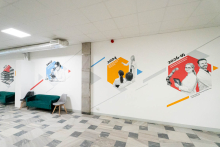 Jasny, szeroki korytarz uczelni z grafikami na ścianach; po prawej widoczny napis ‘100 lat Wydziału Farmaceutycznego WUM 1926–2026’. W głębi kilka osób siedzi i rozmawia