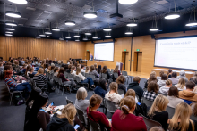 Duża grupa uczestników siedzi w sali konferencyjnej i słucha prezentacji prowadzonej przez prelegenta stojącego przy ekranie, na którym wyświetlany jest slajd dotyczący statystyk.