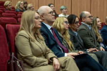 Sześć osób siedzi w jednym rzędzie czerwonych foteli i słucha wystąpienia. Trzymają dłonie na kolanach lub splecione przed sobą. Obok nich siedzą kolejne osoby w dalszych rzędach.