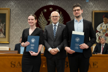 Trzy osoby: kobieta i dwóch mężczyzn, są elegancko ubrani. Kobieta i mężczyzna po prawej trzymają granatowe teczki. W tle siedzą dwie osoby oraz widać duże godło z orłem. 