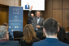 Prelegent stojący na środku sali. Ma uniesioną prawą rękę.  Obok mężczyzny stoi roll‑up z informacjami o konferencji. W tle wisi ekran z widocznym mówcą.