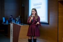 Kobieta w bordowej sukience stoi przy mównicy i przemawia do publiczności, trzymając mikrofon w dłoni. W tle widać fragment slajdu prezentacji wyświetlanej na dużym ekranie.