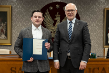 Dwóch mężczyzn stoi obok siebie, są elegancko ubrani. Jeden z nich trzyma teczkę z otwartym dokumentem. W tle siedzą dwie osoby oraz widać duże godło z orłem. 