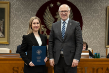 Kobieta i mężczyzna stoją obok siebie, są elegancko ubrani. Kobieta trzyma granatową teczkę. W tle siedzą dwie osoby oraz widać duże godło z orłem. 