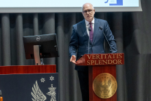 Osoba stoi za mównicą z napisem „Veritatis Splendor” i przemawia do mikrofonów. Trzyma jedną dłoń przy mównicy, a drugą wykonuje gest w powietrzu. W tle widać fragment ekranu z logo UCK WUM.