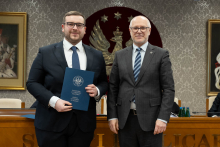 Dwóch mężczyzn stoi obok siebie, są elegancko ubrani. Jeden z nich trzyma teczkę z otwartym dokumentem. W tle siedzą dwie osoby oraz widać duże godło z orłem. 