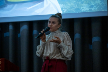 Kobieta stoi przed publicznością i trzyma mikrofon przy ustach. Drugą dłonią wykonuje gest podkreślający wypowiedź. W tle widać fragment wyświetlanego slajdu.