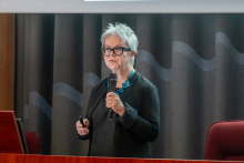 Kobieta stoi przed publicznością i trzyma mikrofon w jednej dłoni, a w drugiej pilot do prezentacji. Kieruje wzrok w stronę słuchaczy i mówi. Za jej plecami widać zasłony oraz fragment ekranu.