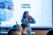 Osoba prowadząca prezentację stoi przed publicznością, trzyma mikrofon i mówi, a za nią na dużym ekranie wyświetlany jest slajd zatytułowany ‘W zderzeniu z Reality Day