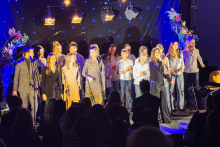 Grupa kilkunastu osób stoi razem na scenie i kończy wspólny występ. Część trzyma mikrofony, a pozostali stoją blisko siebie i patrzą w stronę publiczności. Ich postawa jest swobodna, a gesty sugerują radość i energię po wykonaniu utworu. Widzowie obserwują ich sprzed sceny. 