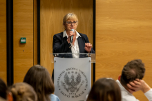 Osoba stojąca za mównicą z logo uczelni przemawia do publiczności, trzymając mikrofon w dłoni w sali wykładowej.