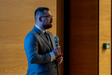 Mężczyzna w eleganckim garniturze stoi przy mównicy i trzyma mikrofon, przygotowując się do wystąpienia w sali konferencyjnej.