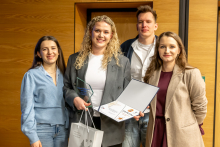 Grupa osób pozuje do wspólnego zdjęcia podczas wydarzenia; jedna z nich trzyma statuetkę oraz otwartą teczkę z dyplomem, a w drugiej ręce ma torbę z upominkami.