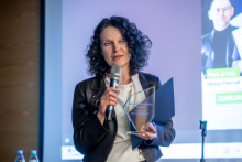 Osoba stojąca na tle ekranu prezentacji trzyma mikrofon oraz szklaną statuetkę, jak podczas wręczenia nagrody lub wyróżnienia