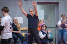 A man standing in the sports hall raises his hands in a gesture of joy, while other participants sit or stand nearby, clapping and watching the situation.