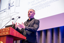 A person in a black suit is standing at a lectern with the inscription ‘Veritatis Splendor’, gesturing while presenting. In the background, a slide with a drawn graphic and the speaker’s contact details is visible.