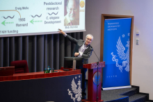 A person standing at a podium and pointing to a presentation slide showing stages of a scientific career and illustrations related to C. elegans research; a blue banner of the Medical University of Warsaw is visible to the side