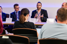 Five people are sitting at a table during a panel discussion. The person in the middle is holding a microphone and speaking. In front of each person lies a nameplate with their first and last name; some of the names are only partially visible. In the foreground, several audience members are visible, seated with their backs to the camera. The background has a light purple tint.
