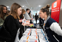 A person providing information at a recruitment booth, talking to visitors and handing out leaflets, with a table arranged with informational materials and a banner reading “Information Recruitment” in the background.