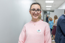 A person in a light pink sweater is standing in a hallway, wearing a badge with a colorful design; in the background, several people are talking near an open doorway