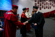 A man in an academic gown shakes hands with one of the three professors standing in front of him. He is holding a document in his other hand and looking toward the professor.