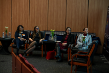 Four people are seated in armchairs arranged in a semicircle, waiting for their part of the event. They rest their hands on their knees or on the armrests. In front of them are small tables with bottles of water and vases with tulips. In the background, wooden panels and a fragment of an informational poster are visible.