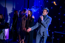 Four people are standing on stage, preparing to sing together. A man with a microphone is standing at the front, making an open‑hand gesture as if introducing the performance. Three women are standing behind him at their microphones and singing.